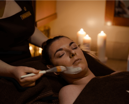 A woman receives a facial treatment with a brush in a dimly lit spa room with candles in the background.