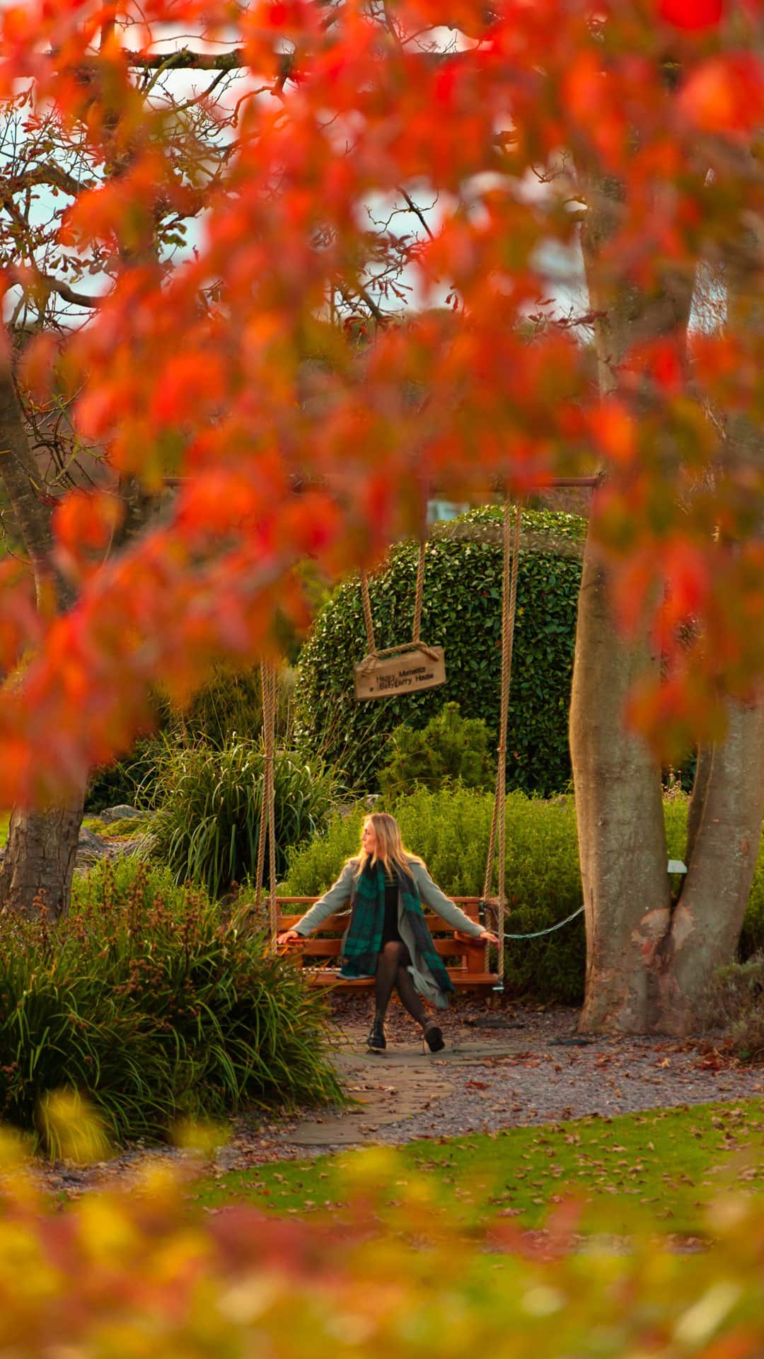 A woman sits on a wooden swing between two trees in a park, surrounded by autumn foliage and greenery.