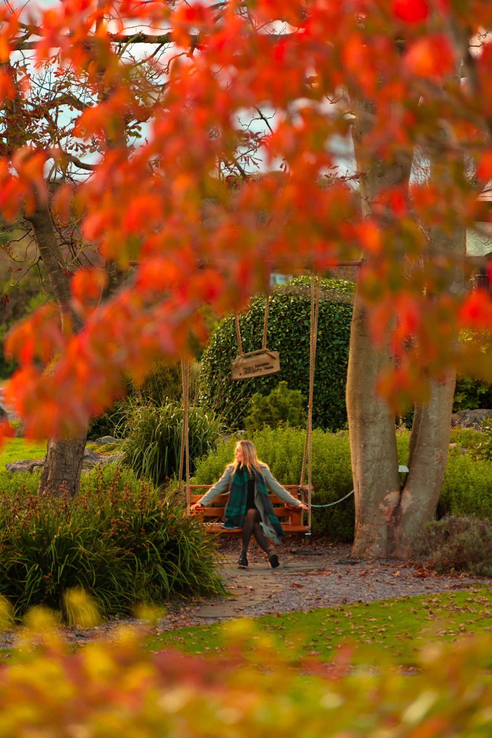 A woman sits on a wooden swing beneath large trees with vibrant red and orange autumn leaves in a garden.
