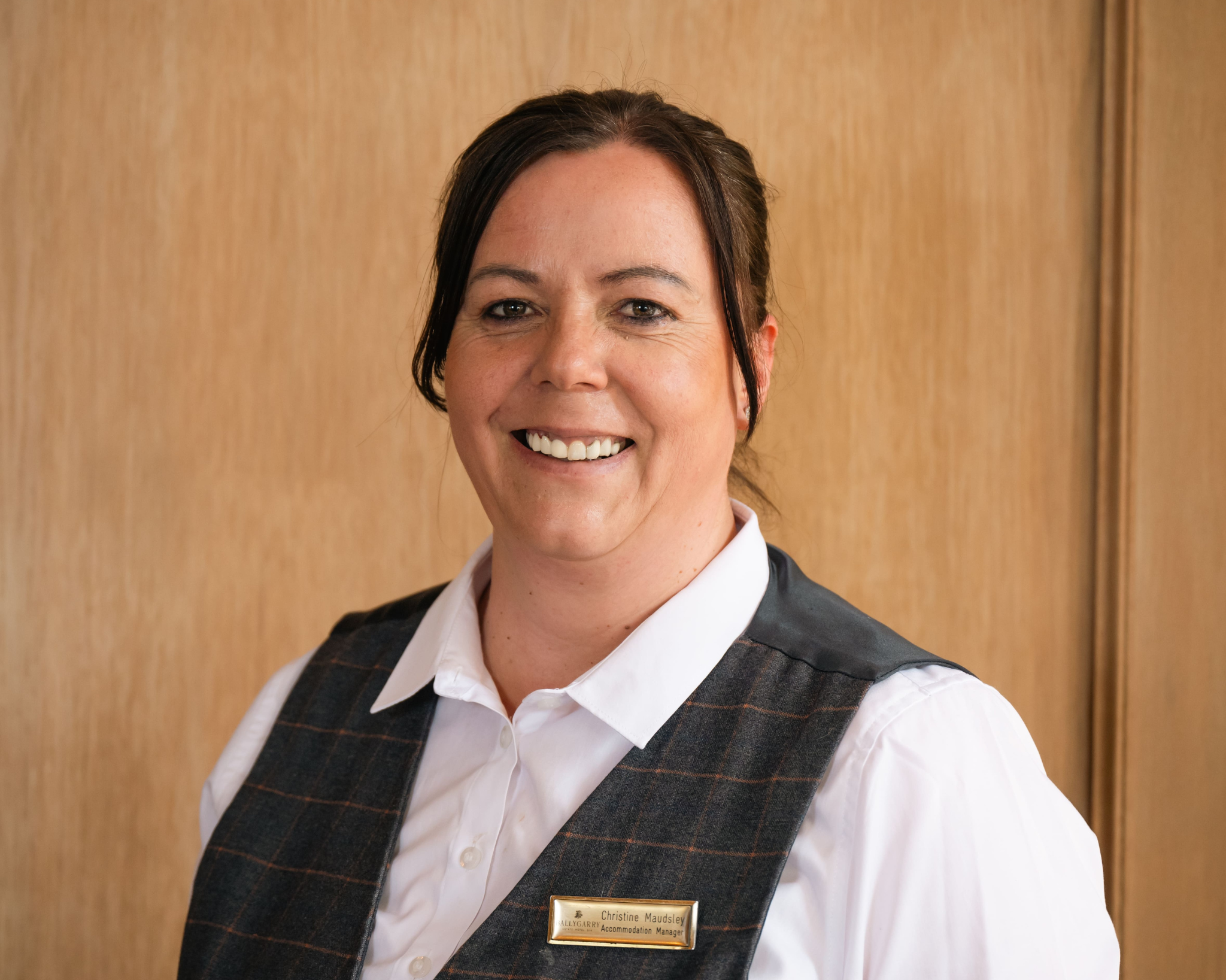 Woman wearing a white shirt and checkered vest, smiling at the camera, standing indoors with a wooden background.