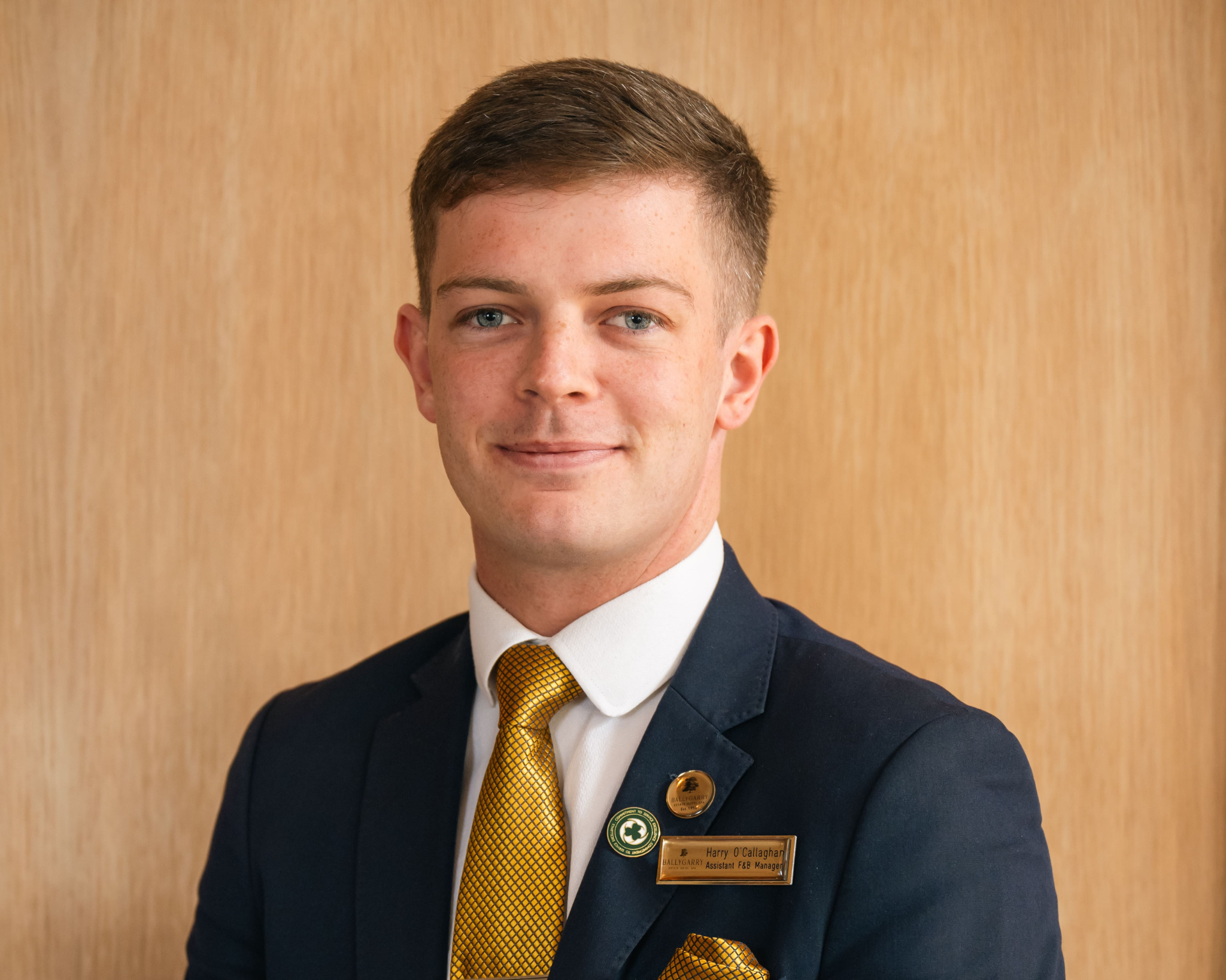 A young man in a dark suit with a gold tie stands in front of a light wood background, wearing a name badge and a lapel pin, and facing the camera with a slight smile.
