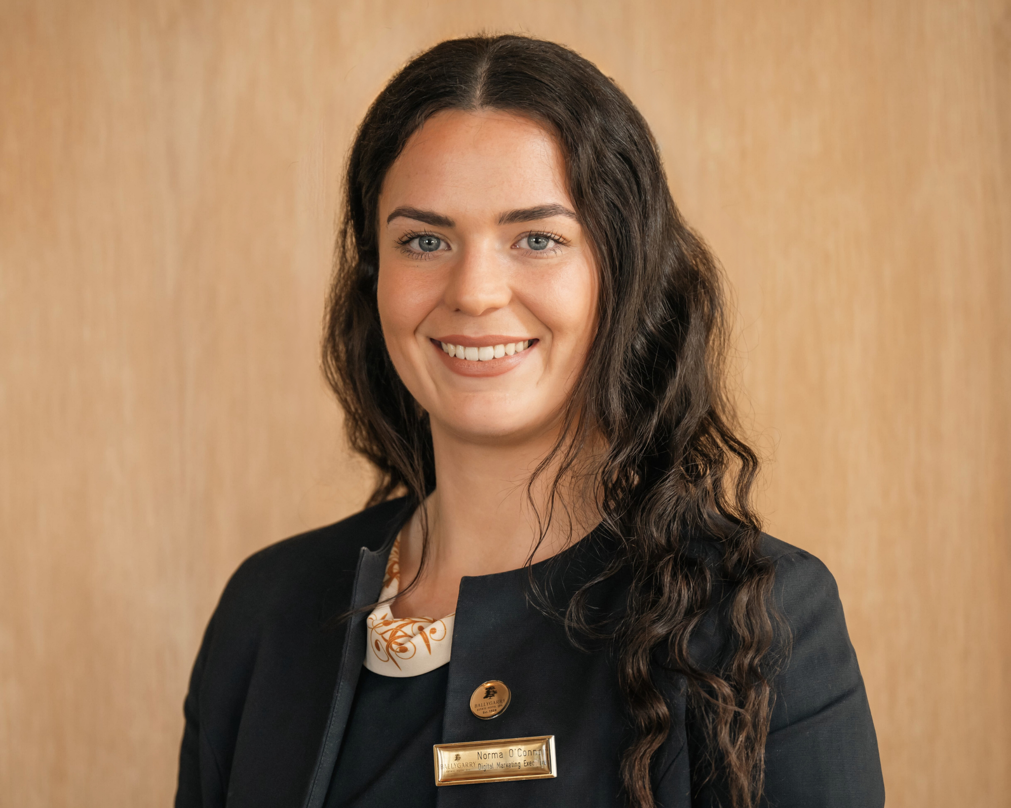 A woman with long, wavy dark hair wearing a dark blazer and a name badge stands in front of a beige background, smiling at the camera.