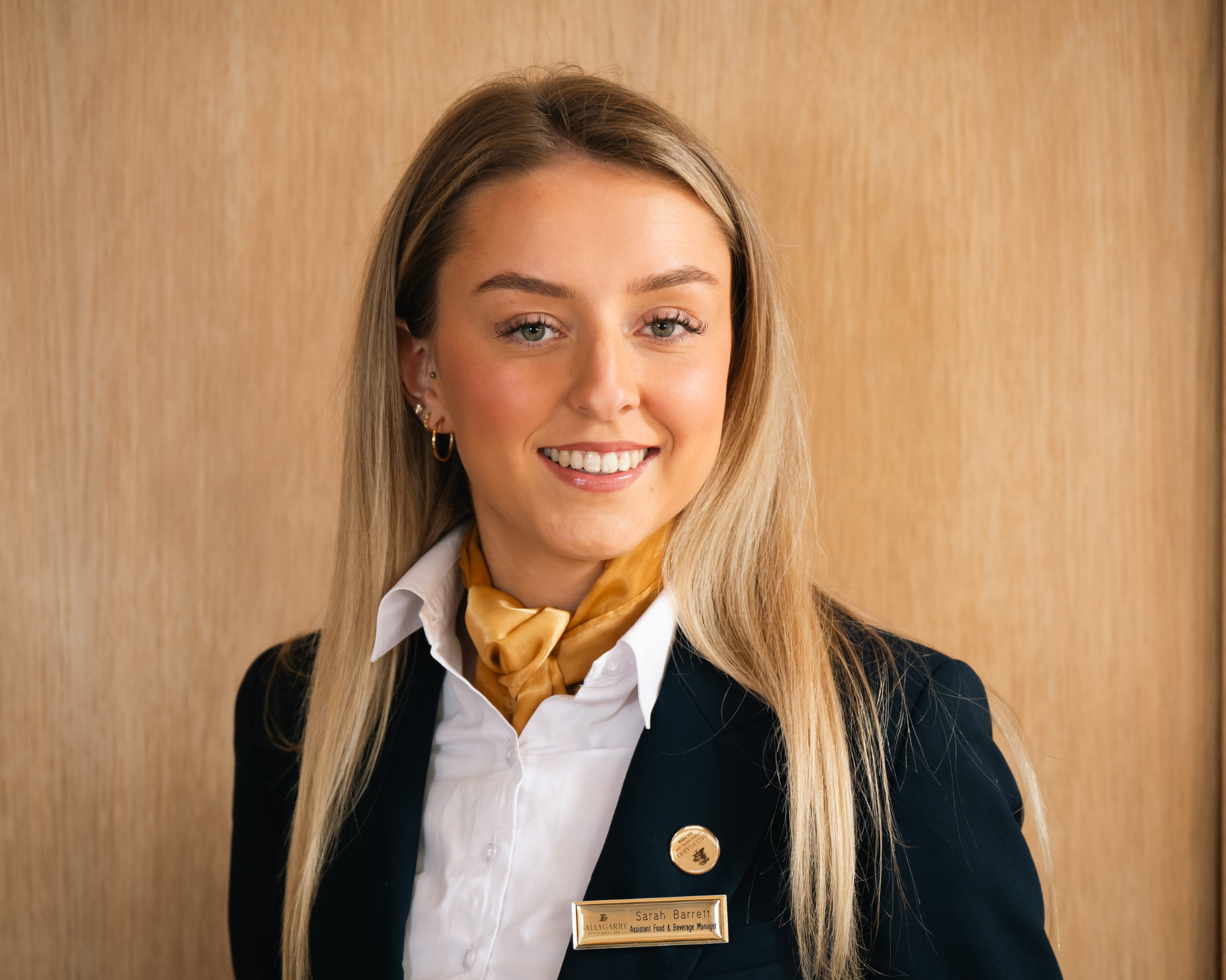 A woman with long blonde hair in a navy blazer, white shirt, and yellow scarf, wearing a name badge, stands in front of a wooden background and smiles at the camera.