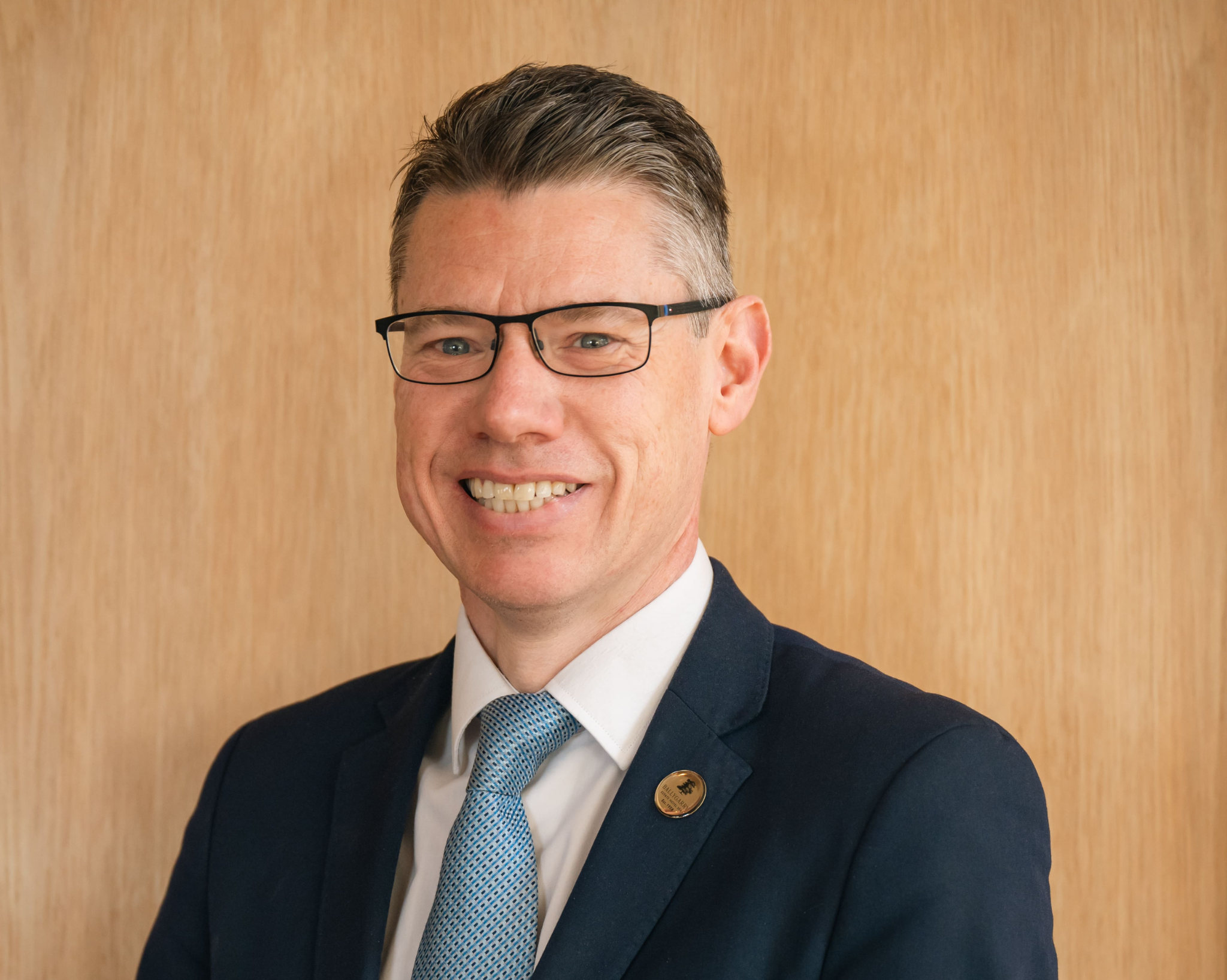 A man wearing glasses, a suit, and a blue tie stands in front of a plain wooden background, smiling at the camera.