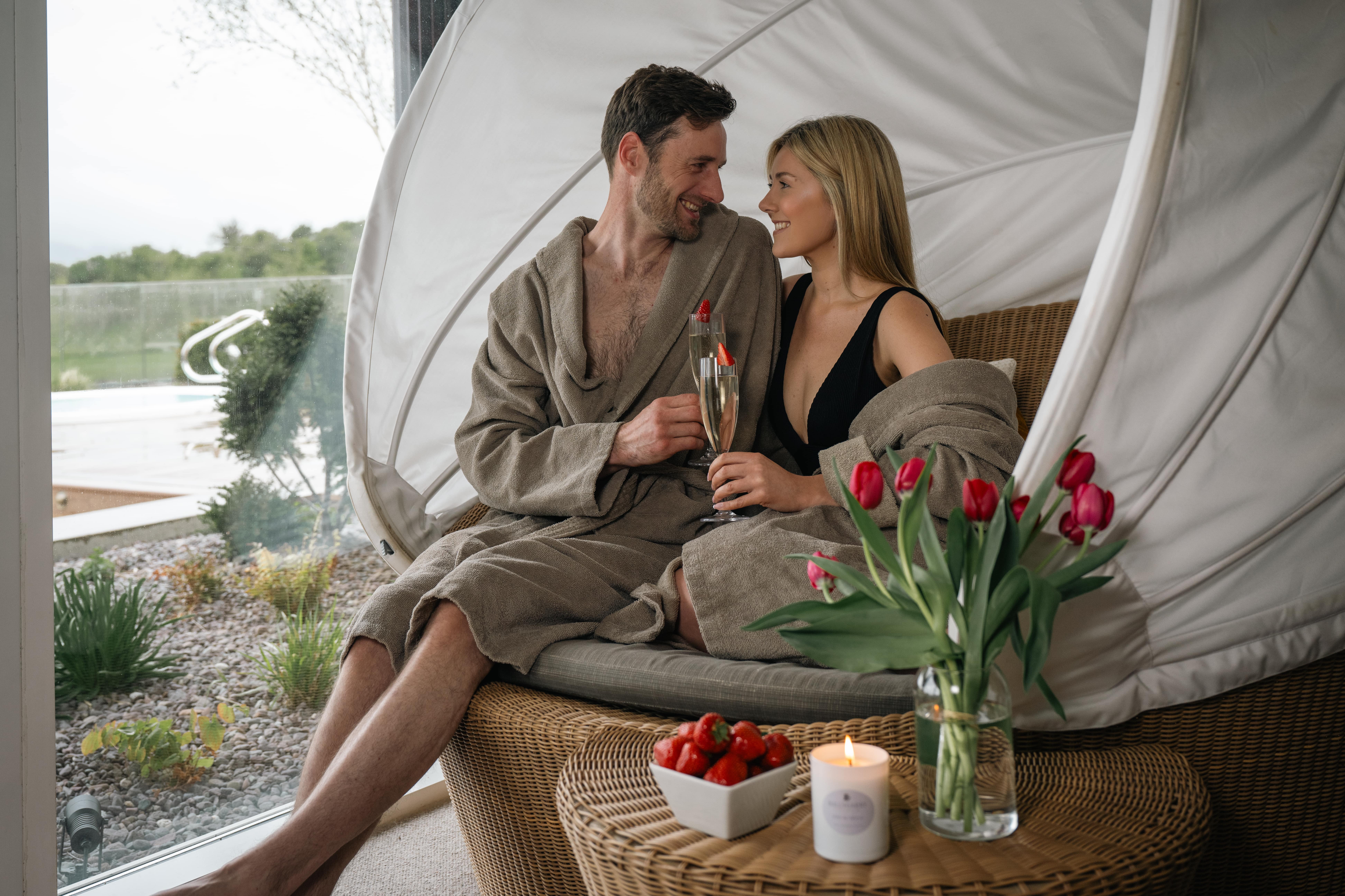 A man and woman in robes sit together on a wicker lounge chair indoors, holding drinks. A vase of red tulips, strawberries, and a lit candle are on a table in the foreground.