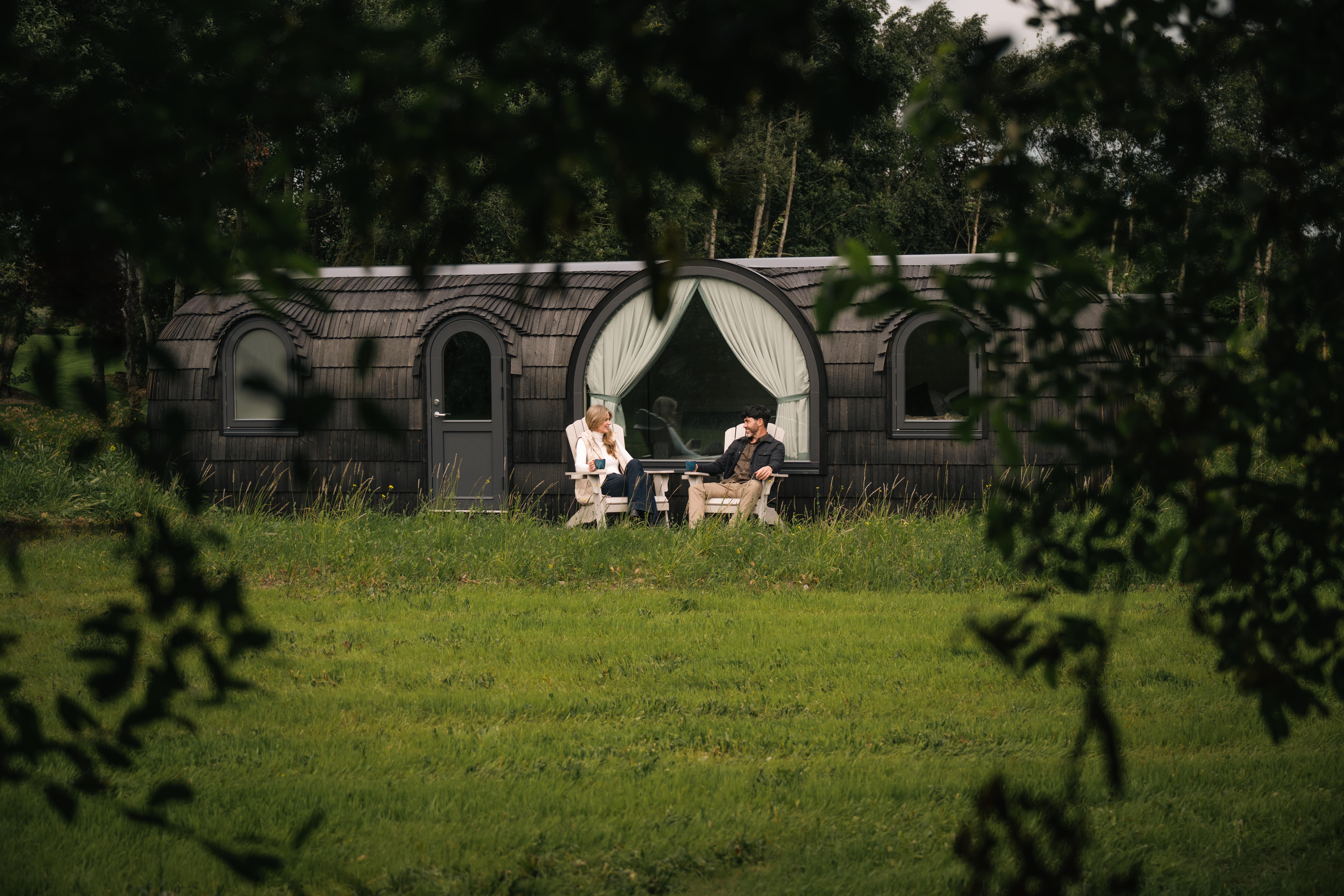 Two people sit on outdoor chairs in front of a black, curved tiny house with large windows, surrounded by green grass and trees.