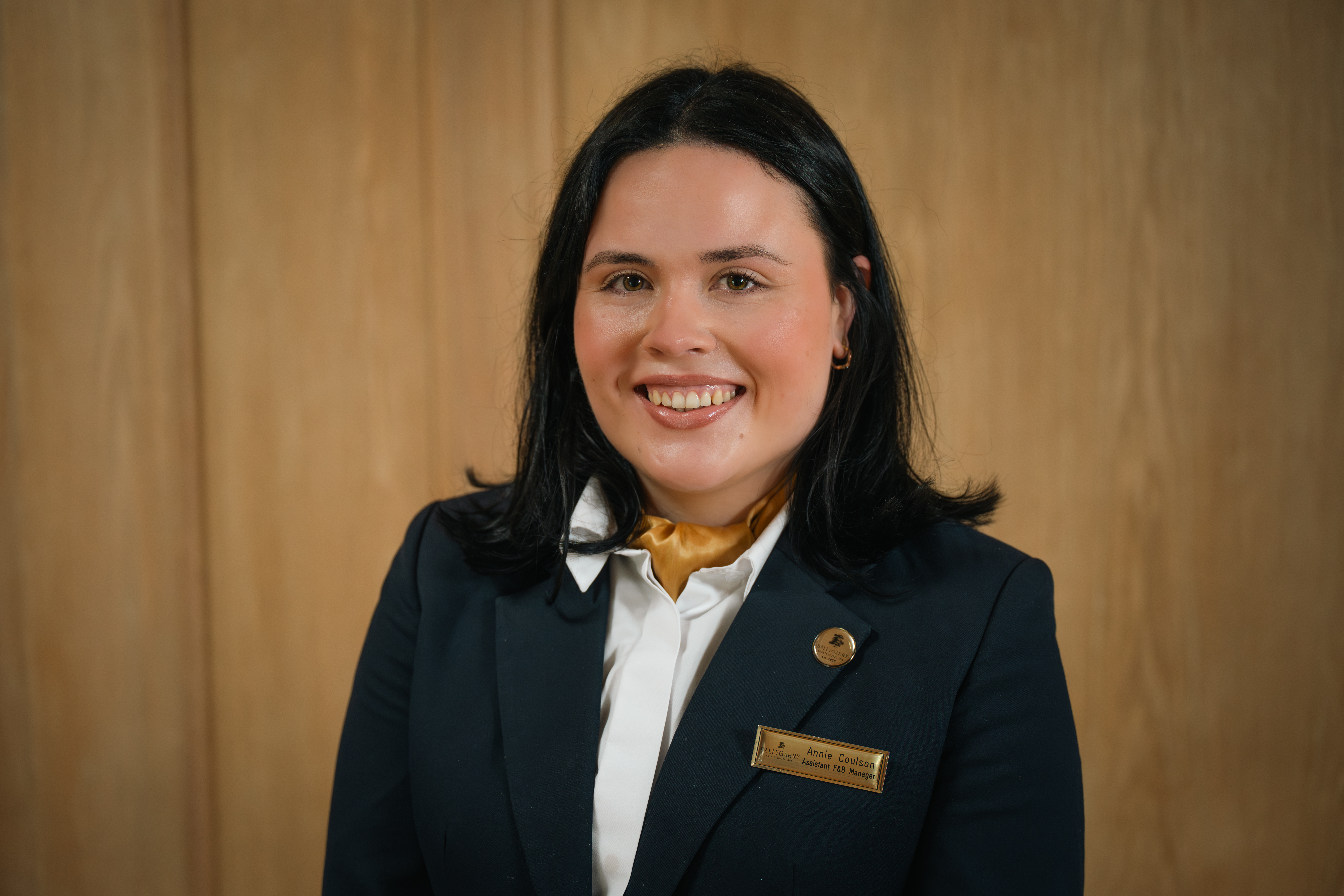 A woman with shoulder-length dark hair, wearing a dark blazer, white shirt, gold scarf, and a name badge, stands in front of a wooden background, smiling at the camera.