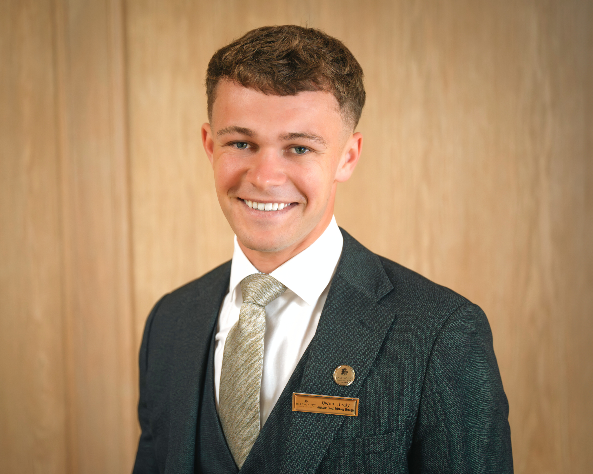 Owen Healy in a suit and tie smiles at the camera, wearing a name tag and standing in front of a plain wooden background.