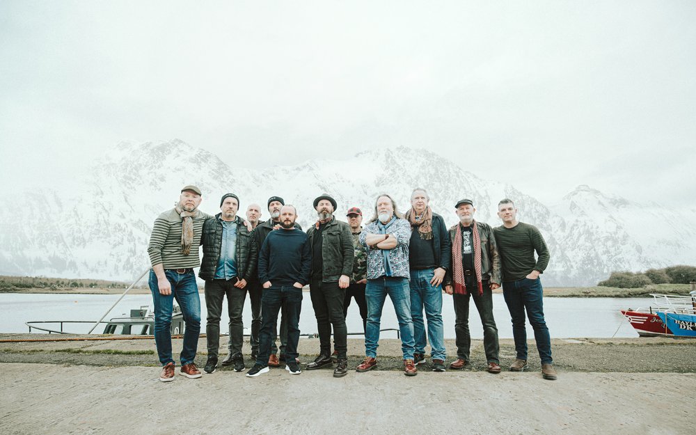 A group of ten men stand side by side outdoors in front of a snowy mountain landscape and a body of water, with boats visible in the background, capturing the serene spirit of a yoga retreat.