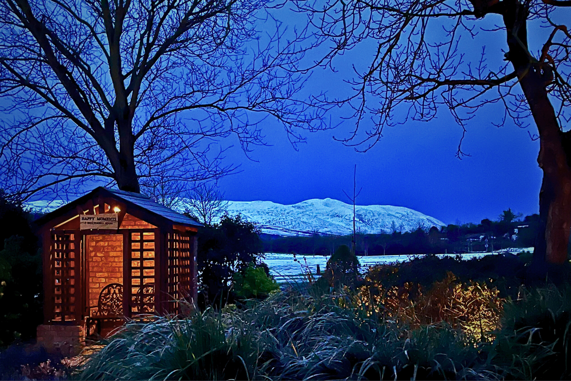 A wooden garden shelter with a bench sits under leafless trees at dusk, with snow-covered hills and a dark blue sky in the background.