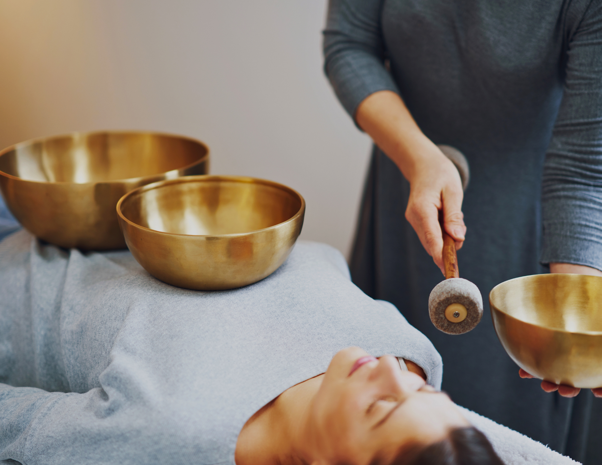 A person lies on their back with three brass singing bowls placed on their body while another person holds a mallet near one of the bowls.