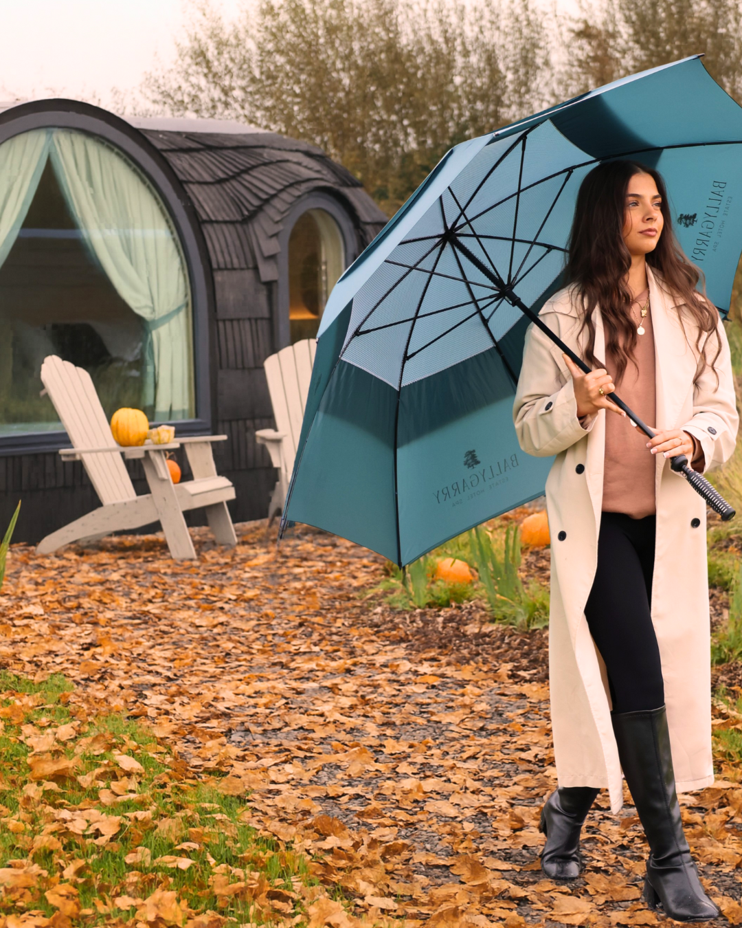 A woman in a long trench coat holds a blue Burberry umbrella, walking on a leaf-covered path near a cabin with pumpkins and Adirondack chairs outside.