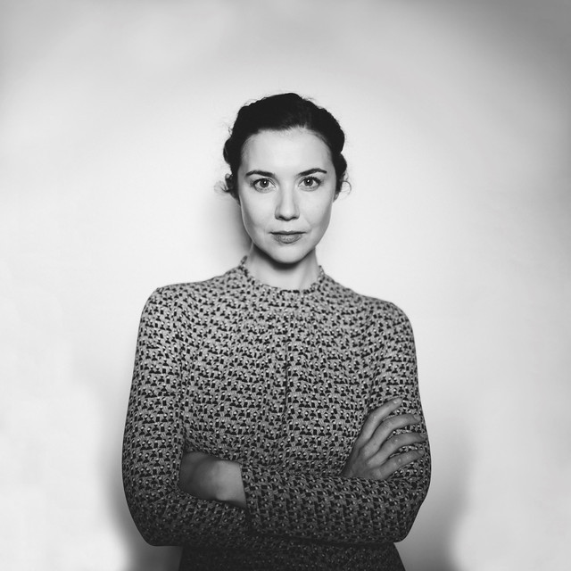 A woman with dark hair pulled back stands with arms crossed, wearing a patterned long-sleeve top, reminiscent of fabrics seen at a Yoga Retreat, against a plain background in a black and white photo.