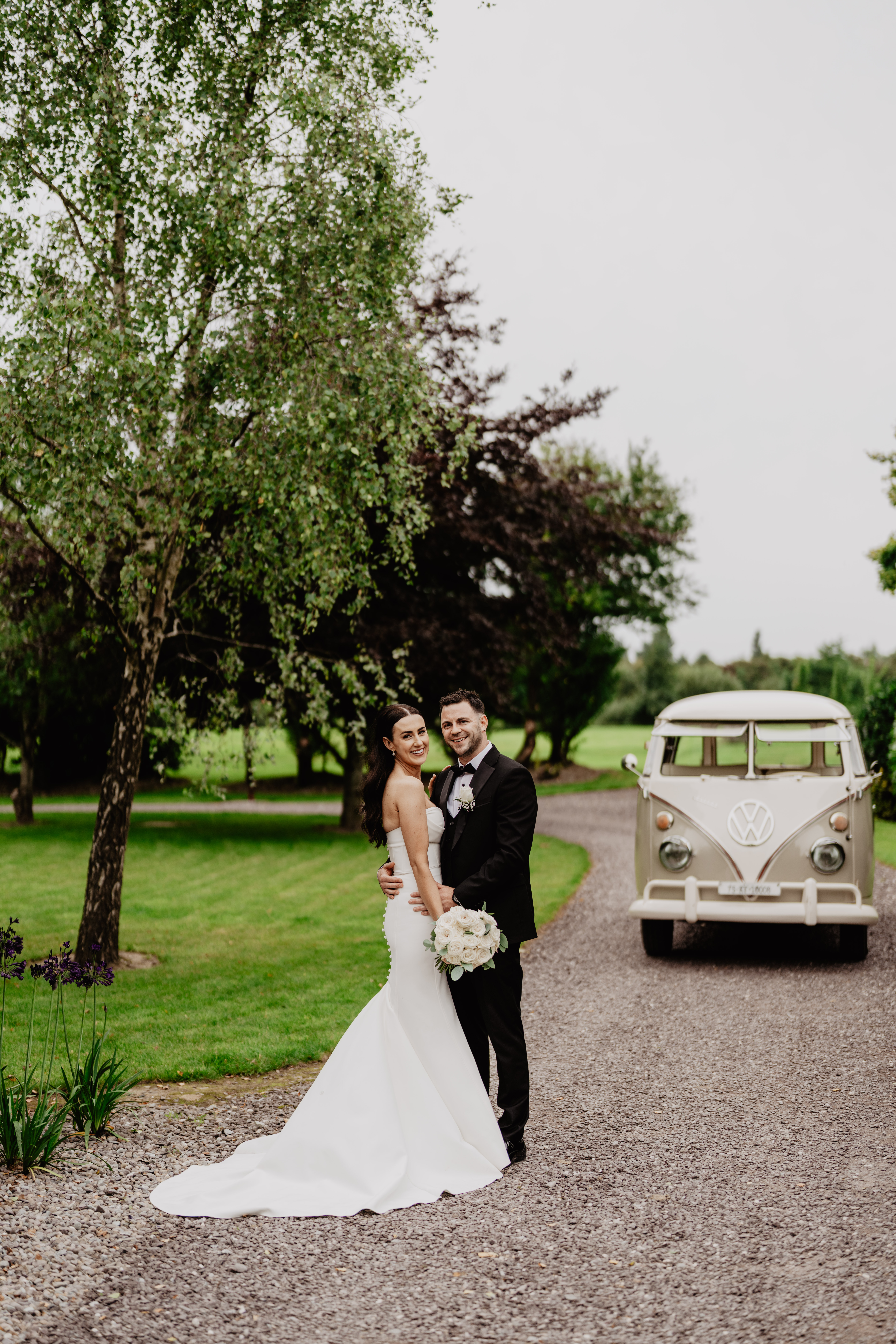 A bride and groom stand together on a gravel path, smiling at the camera, with a vintage Volkswagen van parked nearby and trees in the background.