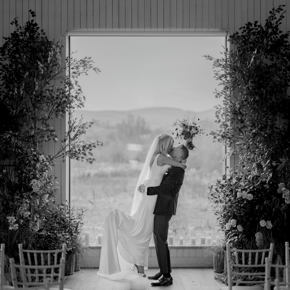 A groom lifts and kisses a bride inside a decorated venue with large windows overlooking a natural landscape.
