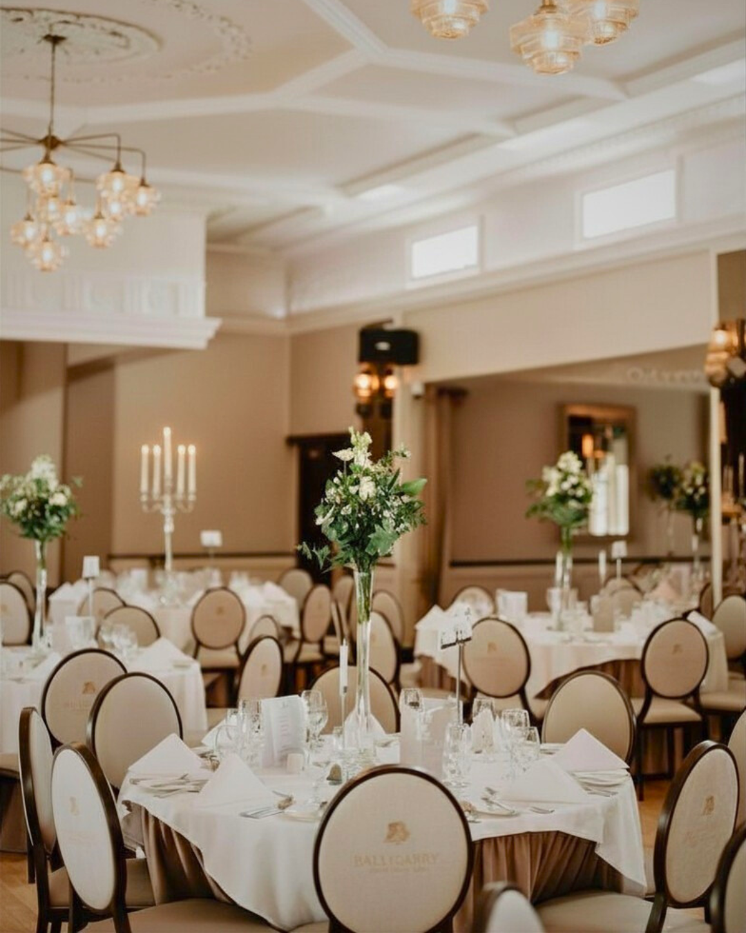 Elegant banquet hall with round tables set for an event, featuring white tablecloths, floral centerpieces, candelabras, and padded beige chairs under chandeliers.