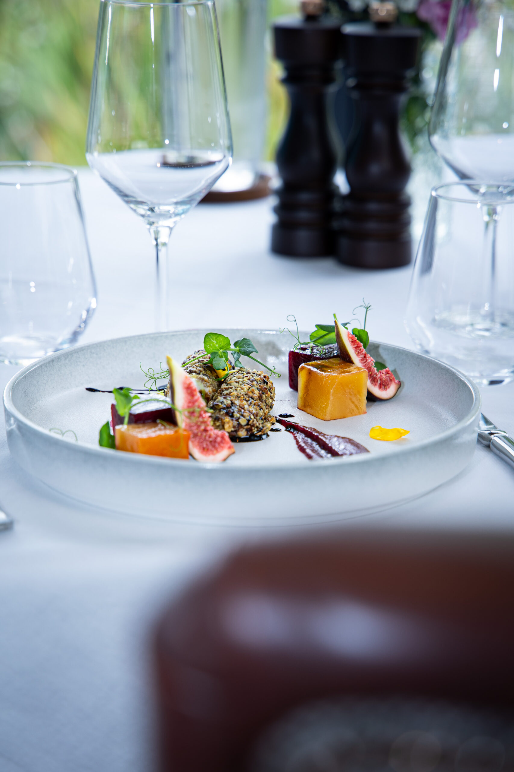 A plated gourmet dish with sliced figs, a round nut-crusted item, orange cubes, microgreens, and sauce, set on a white tablecloth with empty wine glasses and pepper mills in the background.