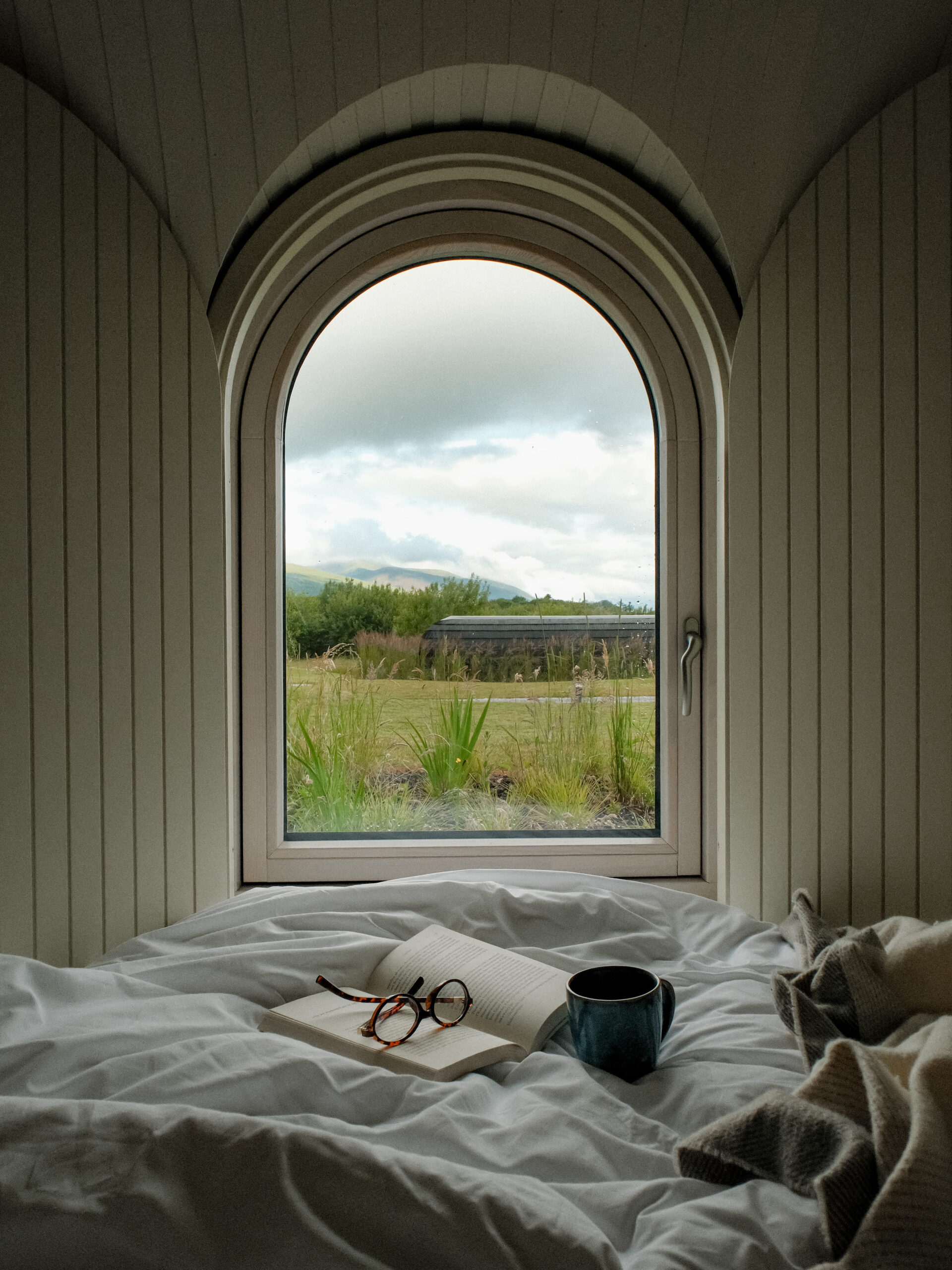 An arched window at the Estate Cabins at Ballygarry overlooks greenery and sky, with a bed below holding an open book, eyeglasses, and a mug.